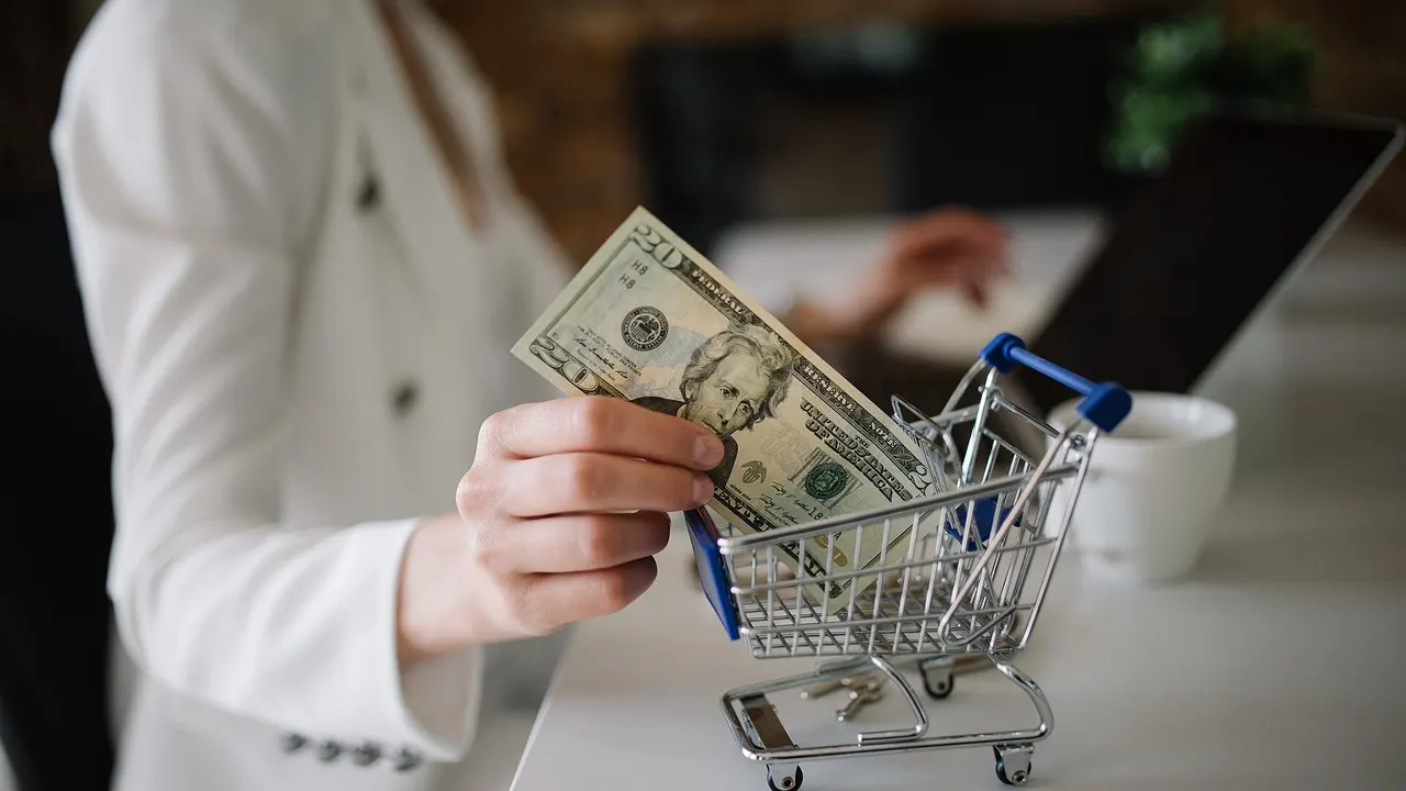 A woman at a desk shopping online with a small toy shopping cart and cash beside the laptop